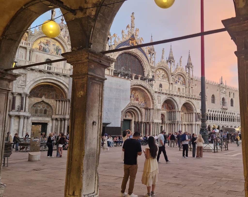 View of St. Mark’s Basilica from the Procuratie at sunset, with mosaics, arches and visitors in Piazza San Marco.