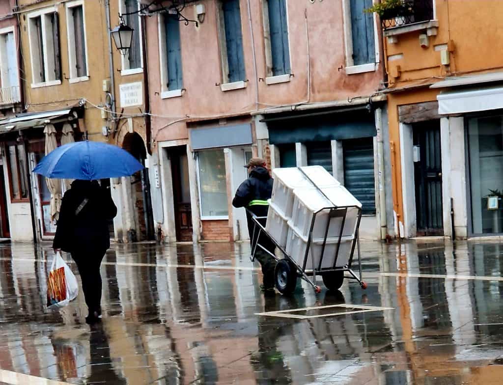 Worker pulling a loaded delivery cart through a rainy Venetian street after unloading goods from a boat.